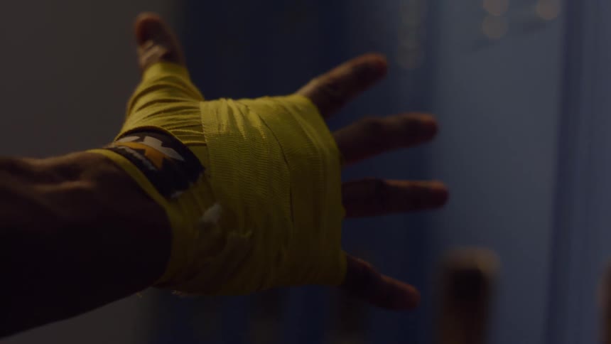 Close-up shot of a boxer's hand wrapped in yellow tape flexing and unflexing their fingers in a dark room