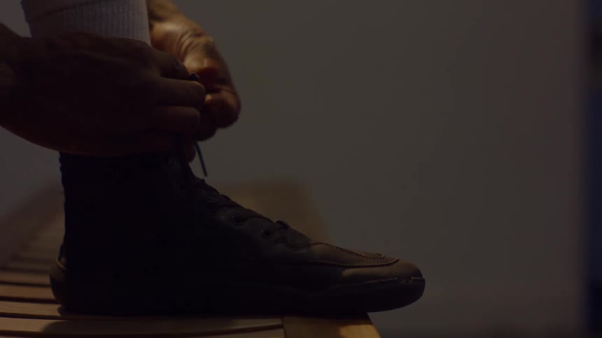 A person's hands are shown in close-up tying the laces of a black high-top shoe in a dark, moody setting
