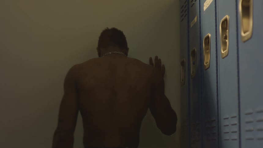 A muscular and shirtless man expresses his frustration and defeat by punching a wall in a dimly lit locker room