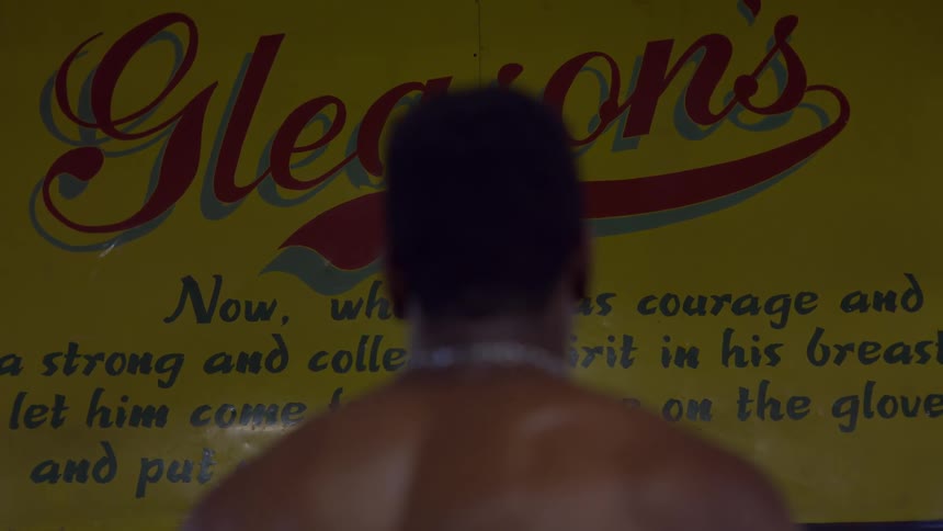 A muscular boxer stands with his back to the camera, facing the iconic Gleason's Gym sign in a moment of quiet determination