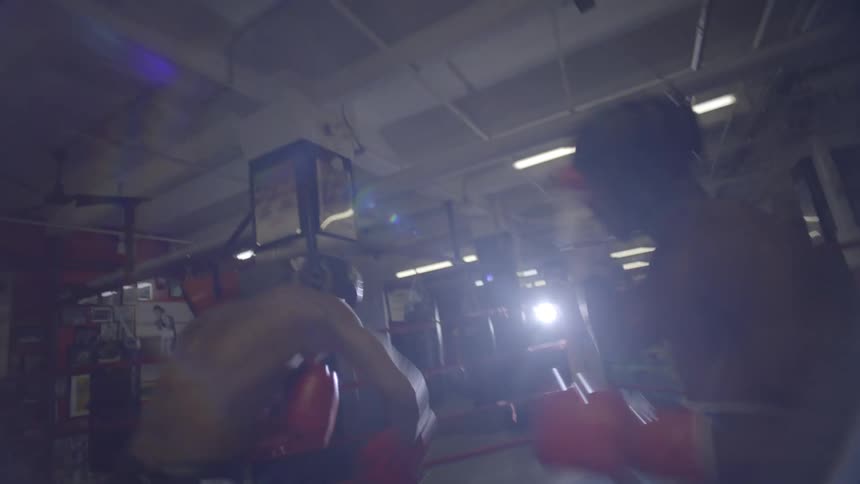 A dramatic point-of-view shot shows two boxers sparring in slow motion inside a gritty boxing ring