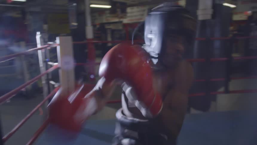 A determined young boxer in headgear and red gloves throws punches at the camera while training in a dimly lit boxing ring