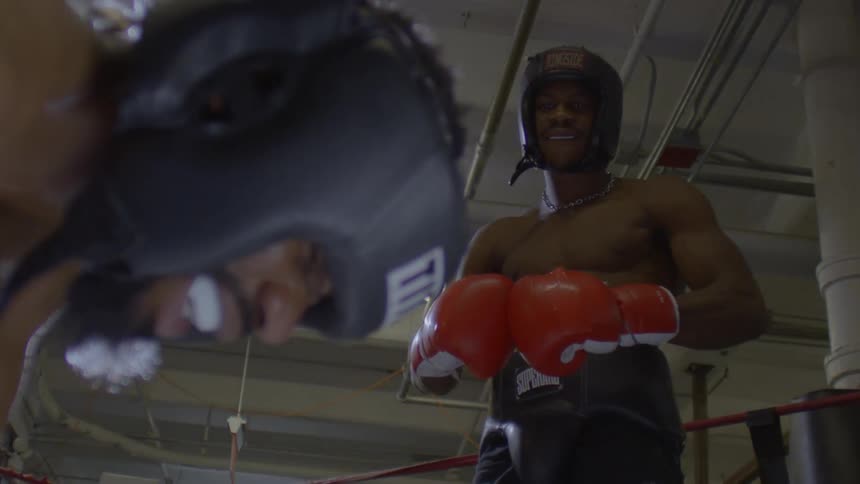 A slow-motion, low-angle view of a shirtless boxer in headgear smiling at his opponent in the ring