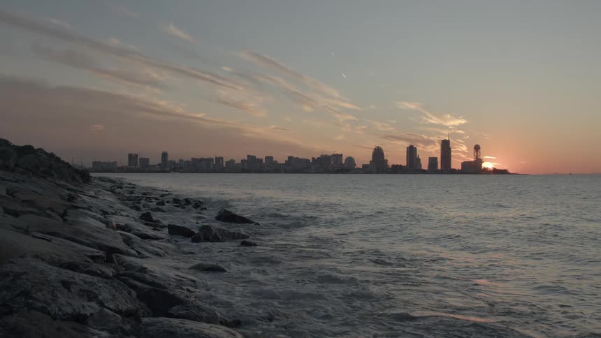 Gentle ocean waves lap against a rocky shore with a silhouetted city skyline during a beautiful sunset in slow motion