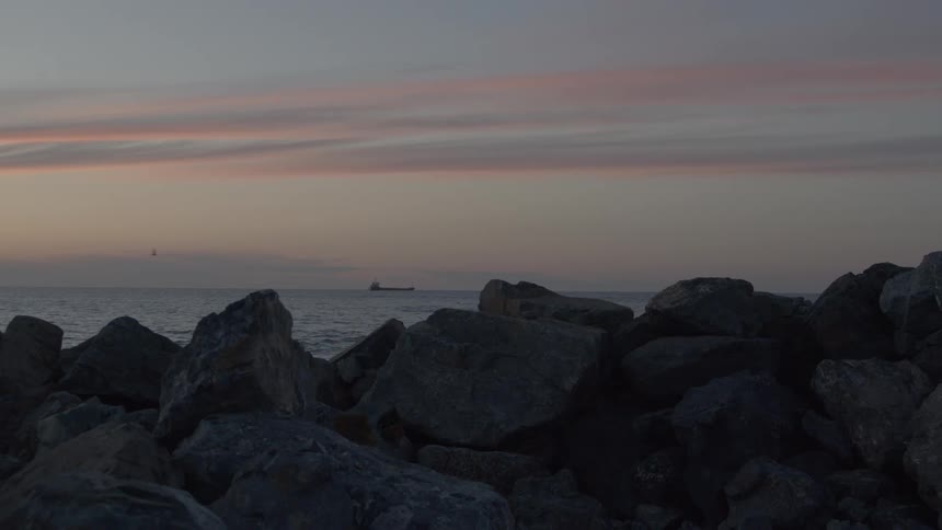 A distant cargo ship sails across the calm ocean under a pastel sunset sky, viewed from a rocky shoreline
