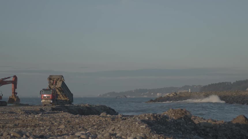 Slow motion captures a dump truck unloading rocks onto a beach during a coastal reinforcement project as seagulls fly by