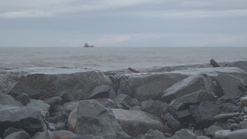 An ocean wave splashes over a rocky breakwater in dramatic slow motion as a ship sits on the horizon