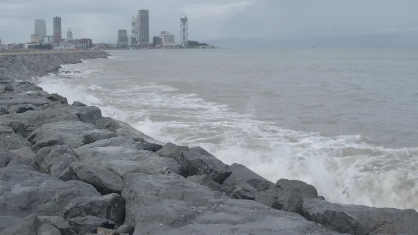 Rough ocean waves crash in slow motion against a rocky seawall with the city of Batumi, Georgia in the background