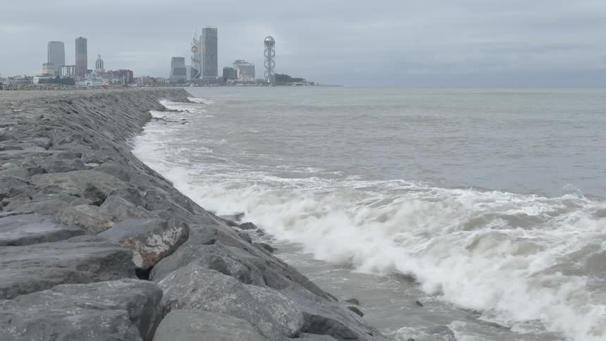 Powerful ocean waves crash in slow motion against a rocky breakwater with a modern city skyline visible in the distance on an overcast day