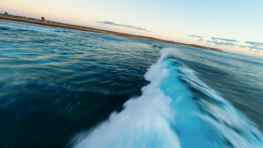 An immersive low-angle drone shot flies alongside a rolling ocean wave as it crests and breaks towards the shore at dawn