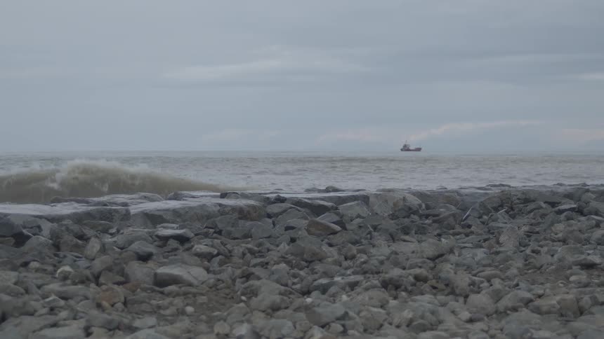 Slow motion footage of powerful ocean waves crashing against a rocky shore with a cargo ship in the distance on an overcast day