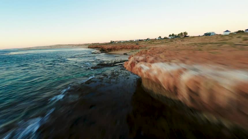A thrilling FPV drone flight speeds along a rugged red rock coastline next to the clear ocean at sunset