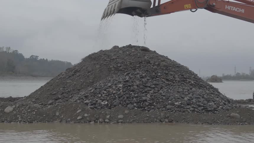 An industrial excavator arm dumps a bucket of wet gravel and sand onto a large pile in a river