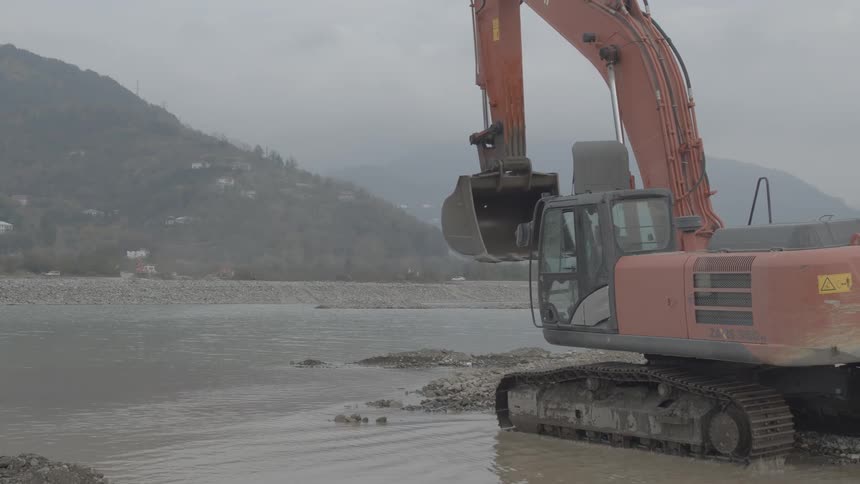 A large industrial excavator maneuvers on the rocky shore of a river in a mountainous region on a cloudy day