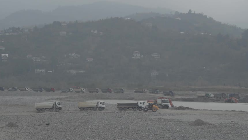 A fleet of dump trucks and excavators work in a large gravel quarry on a hazy, overcast day