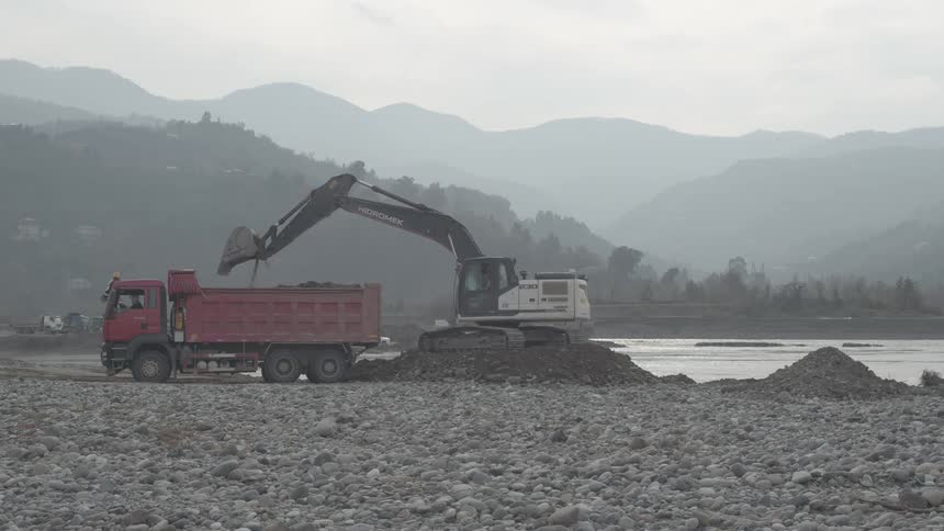 An excavator scoops gravel and soil from a riverbed into a large dump truck in a wide, hazy mountain valley