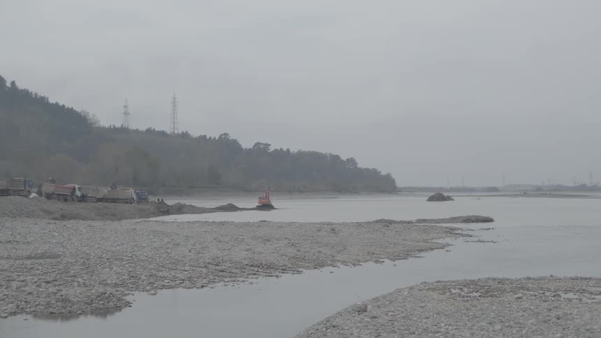 An excavator dredges a rocky riverbed on an industrial site during an overcast and foggy day