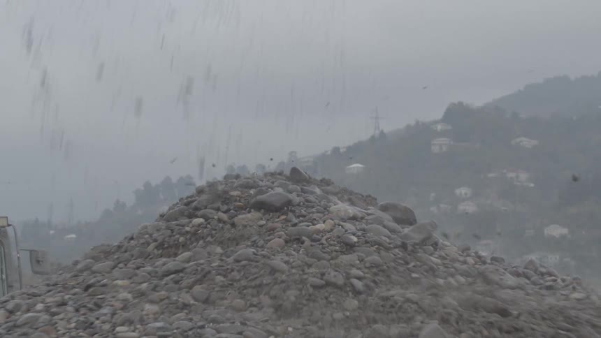 An industrial excavator scoops wet gravel from a riverbed and deposits it onto a large pile on the bank in slow motion