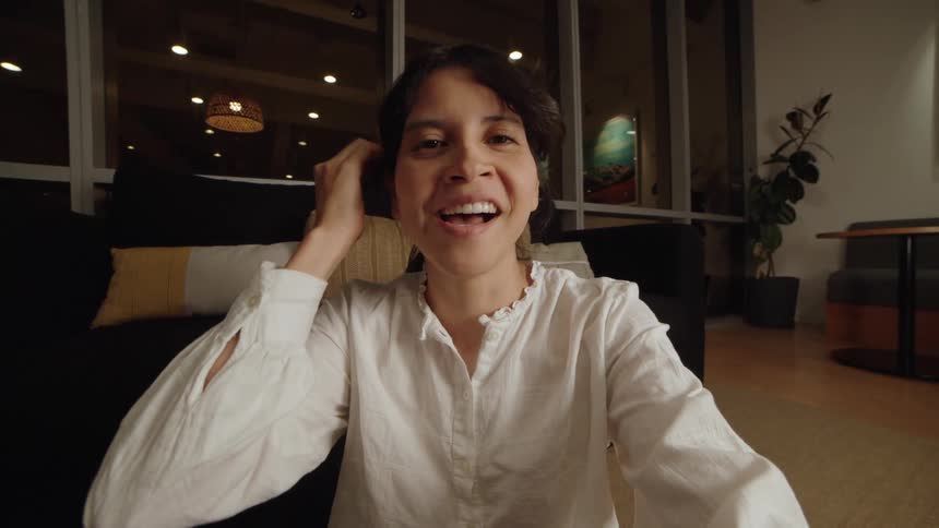 A happy woman smiles and waves while on a video call in her modern living room at night