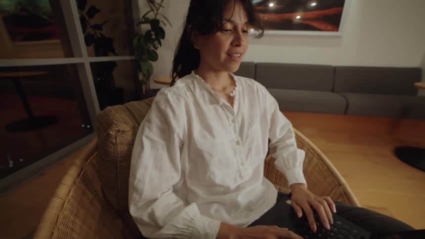 Dolly-in shot of a young woman smiling as she works on her laptop in a comfortable chair in a modern office at night