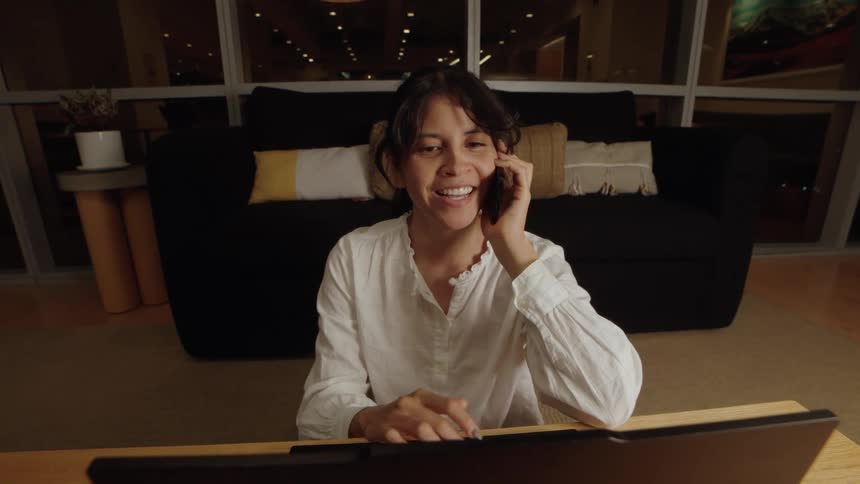 Slow dolly out reveals a happy woman laughing while talking on the phone in front of her laptop at night in a modern apartment