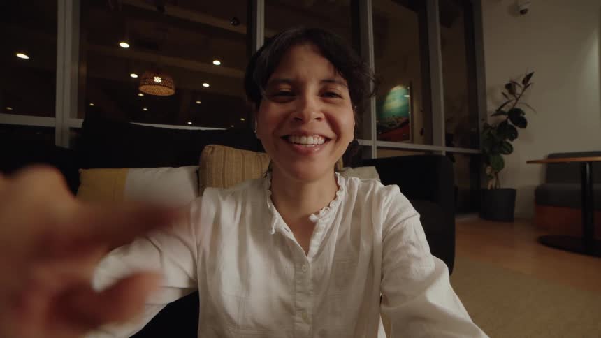 POV of a video call with a smiling woman who playfully interacts with the screen in a cozy, modern room at night