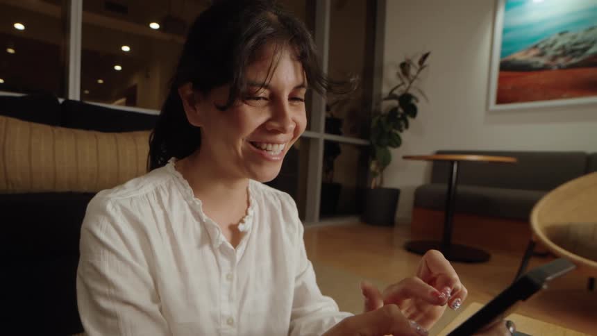 A smiling woman engages in an animated video call on her smart device in a modern lounge at night