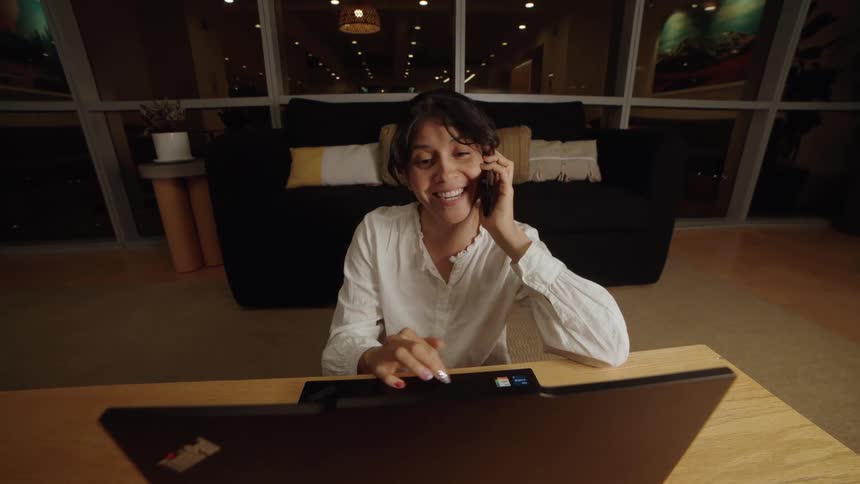 A smiling woman talks on her smartphone while using her laptop, sitting on the floor of a modern living room at night