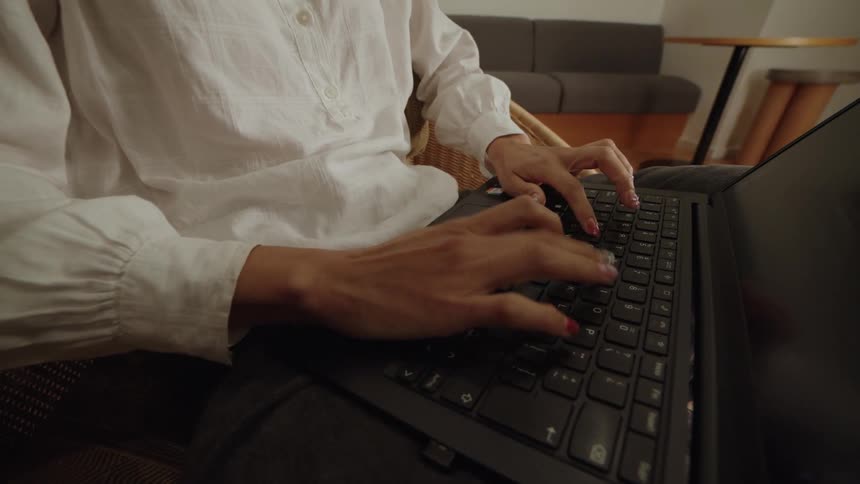 A slow push-in shot focuses on a person's hands as they type on a laptop from a comfortable wicker chair