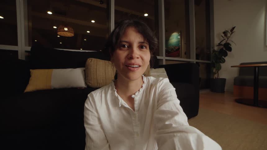 A woman smiles warmly and talks directly into the camera during a video call in her cozy living room at night