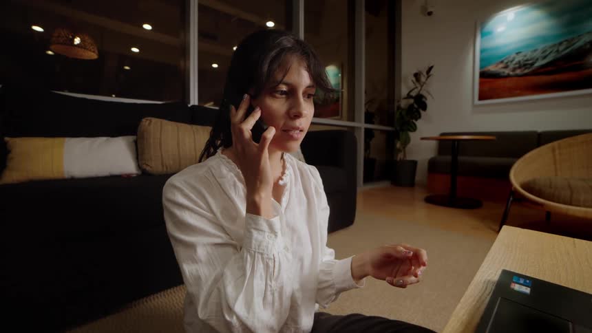 A serious woman answers her smartphone while working late at night on a laptop in a modern office