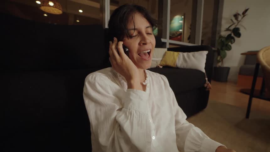 A happy woman working late from her living room smiles and laughs while talking on her phone and using a laptop