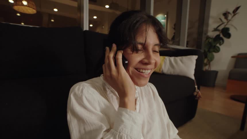 Close-up of a happy woman smiling while having a pleasant conversation on her phone in a cozy living room at night