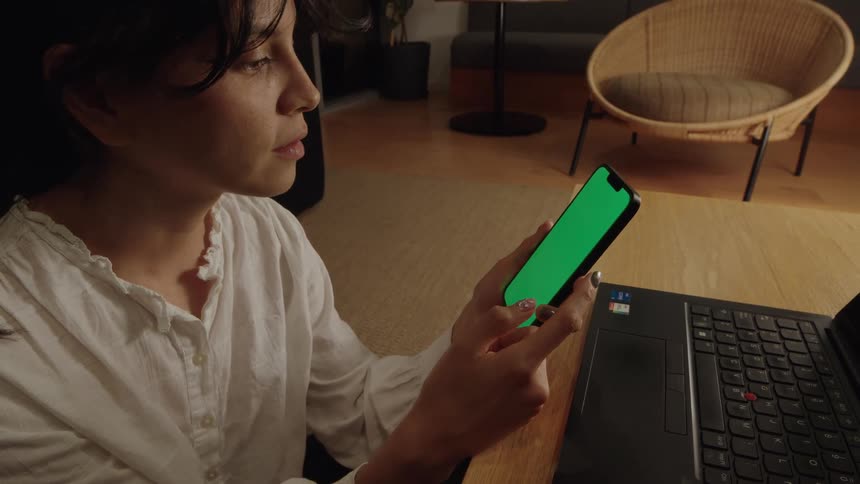 A woman sitting at her desk at night scrolls on her smartphone with a green screen next to her laptop