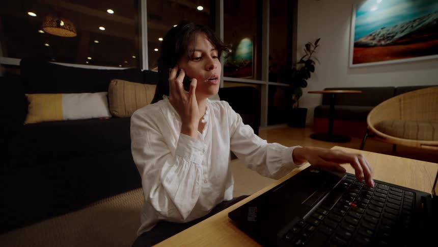 A focused businesswoman works late at night in a modern office, multitasking on her laptop while on a phone call