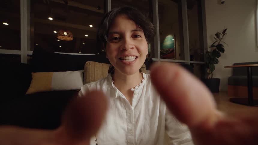 Happy young woman engages in a video call, smiling and talking directly to the camera from a cozy, modern room at night