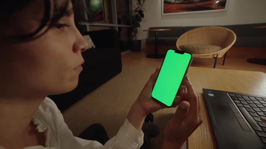 A woman scrolls on her smartphone with a green screen while sitting in her modern living room at night