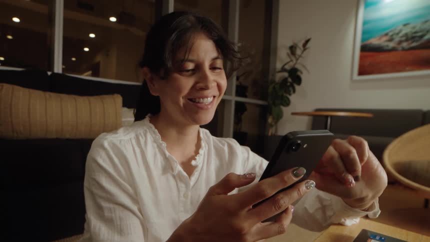 A joyful woman smiles while scrolling through her smartphone in a modern, warmly lit lounge area at night