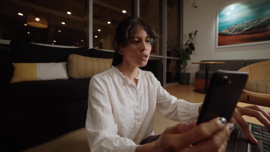 A focused businesswoman participates in a video conference on her smartphone while working on her laptop in a contemporary office at night