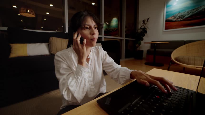 A professional woman talks on her phone and uses her laptop while working late in a modern living room