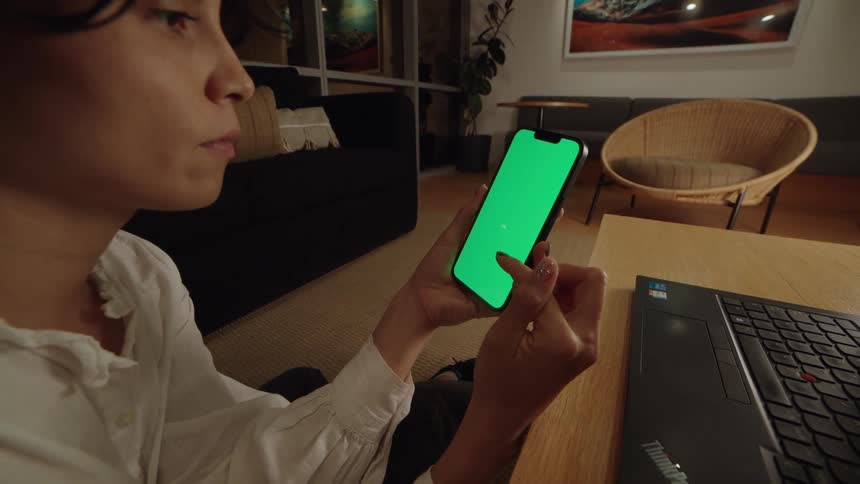 Behind-the-scenes dolly out shot of a woman scrolling on a green screen smartphone in a modern living room set