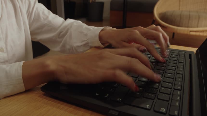 A close-up of a person's hands multitasking, first using a trackpad and holding a phone, then putting the phone down to type on a laptop keyboard