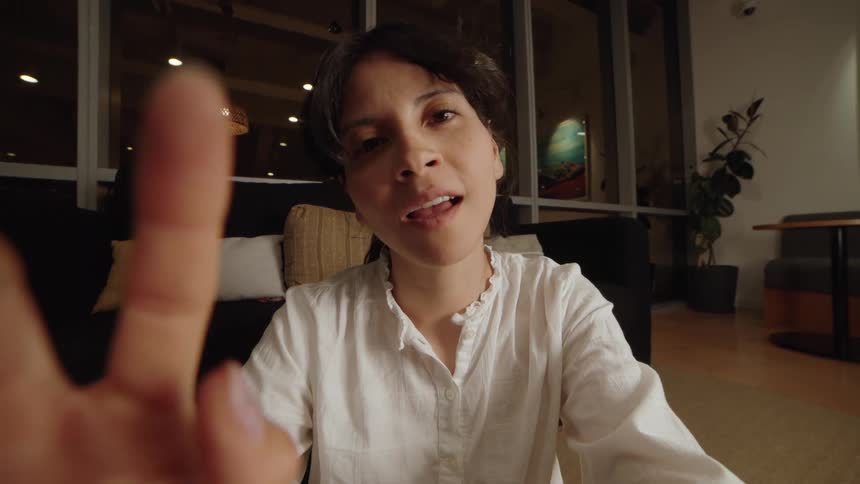 A woman has a shocked reaction while on a video call, looking directly into the camera in a modern living room at night