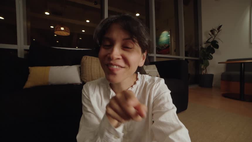An expressive woman shares a story directly with the camera in a cozy, modern room at night