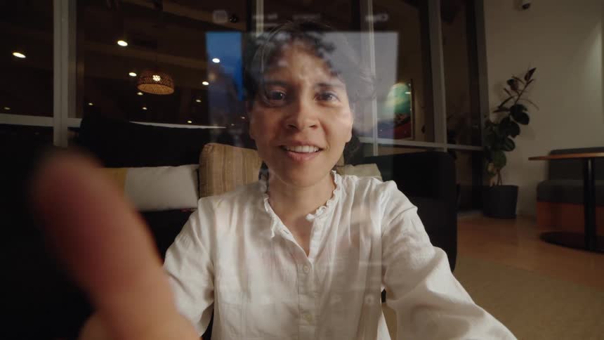 A POV shot of a woman happily using a transparent, futuristic touch screen tablet to scroll through a social media feed in her living room