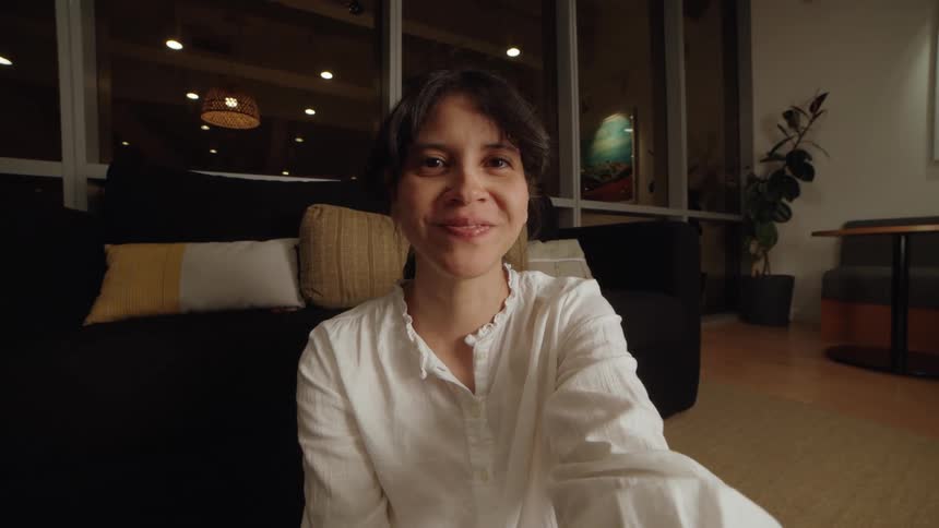 A point-of-view shot of a woman smiling and talking during a video call in her cozy living room at night