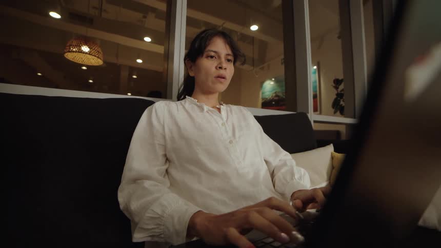 A low-angle view shows a focused woman working late at night, typing on her laptop while sitting on a sofa in a modern office