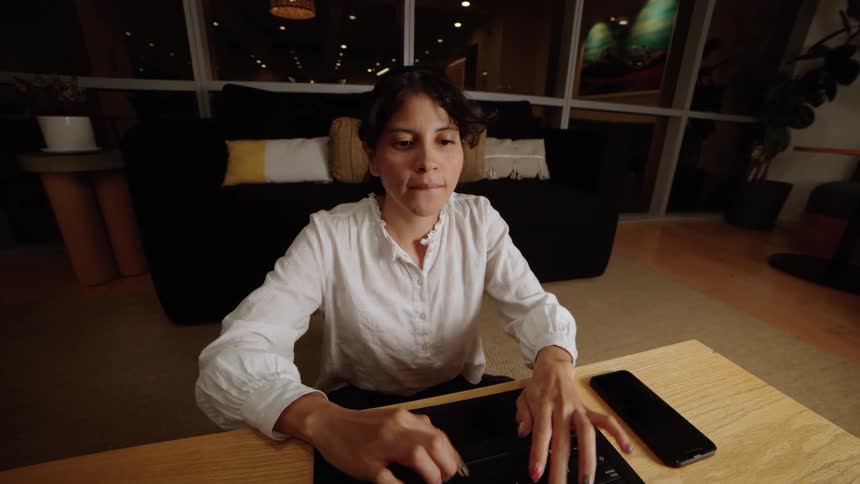A woman sits on the floor at a coffee table, typing on her laptop late in the evening in her modern home