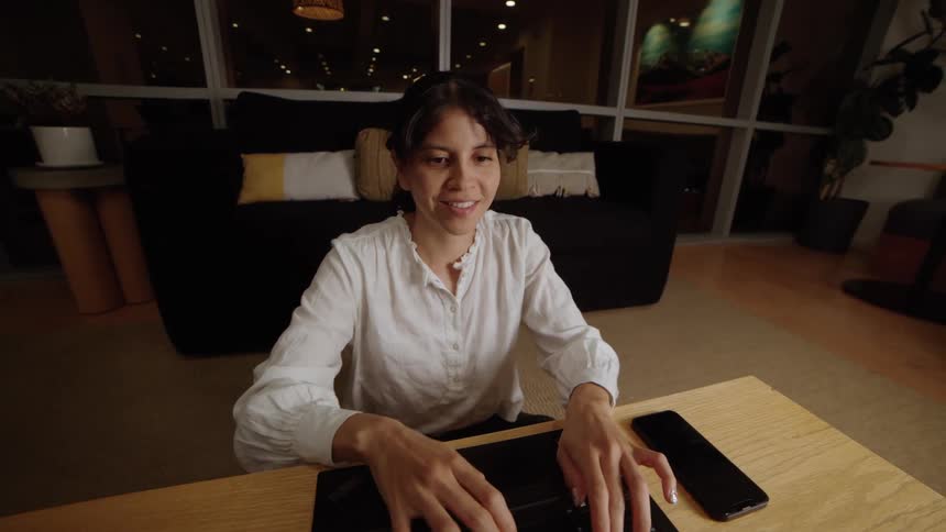 A woman smiles while typing on her laptop, working from her cozy, modern living room at night