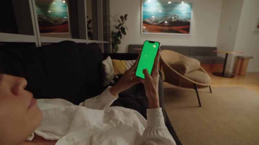 A person transitions from sitting to lying on a couch at night while scrolling on a smartphone with a green screen